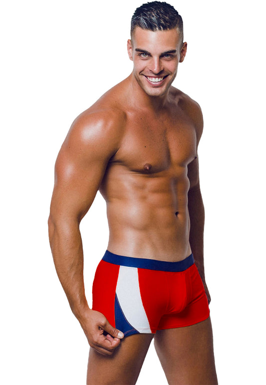 A smiling, muscular man stands confidently in a pair of red, white, and blue Santa Monica Beach Trunks by BWET SWIMWEAR. He has short, dark hair and a well-defined physique. His right hand is pulling slightly on the waistband of the UV-protected fabric trunks, revealing part of his hip. The background is plain white.