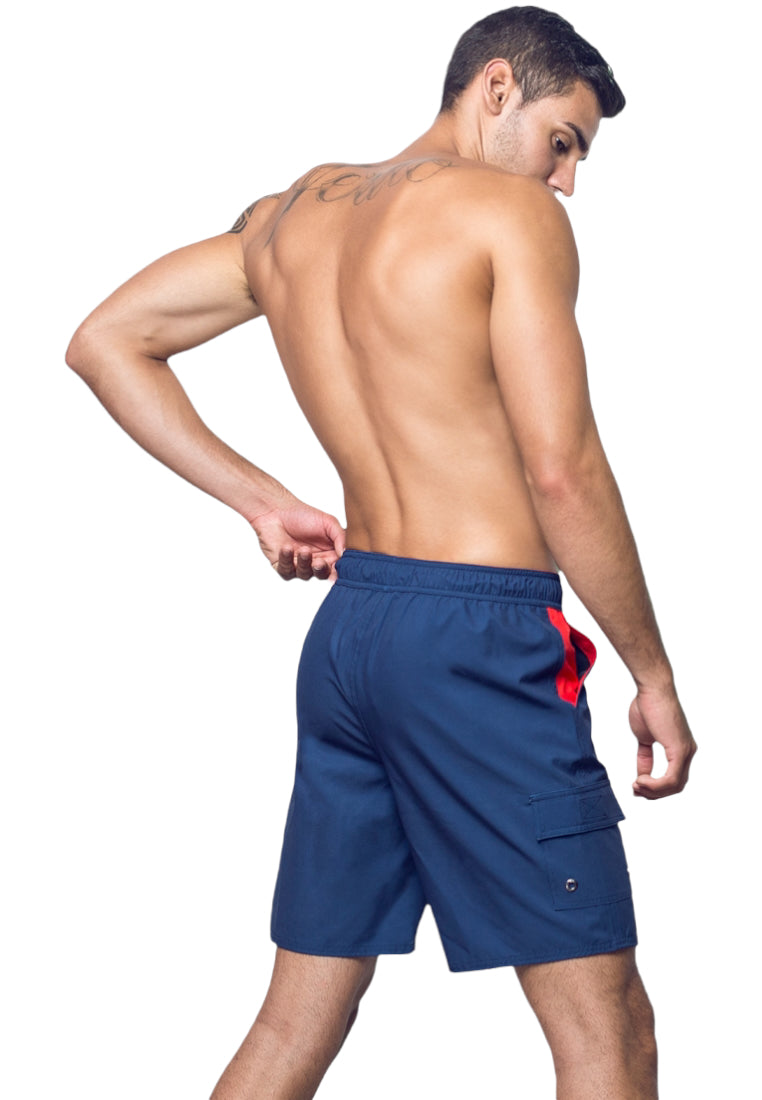 A shirtless man is facing away, showcasing his back and wearing BWET SWIMWEAR's Neptune Beach Shorts, which are blue with red accents and made from recycled material. He has a tattoo on his upper back and appears to be adjusting the waistband of his UV protection shorts. The background is plain and white.