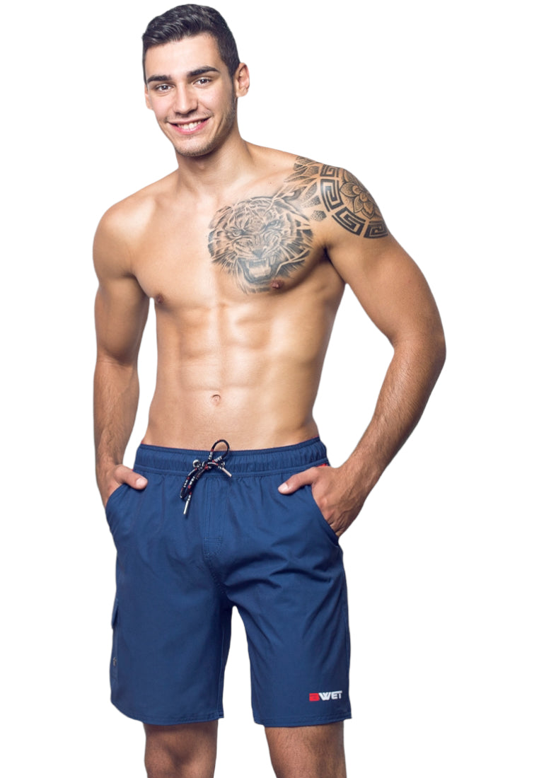 A man with short dark hair is smiling and standing with his hands in the pockets of his BWET Swimwear Neptune Beach Shorts. He is shirtless, displaying a large tattoo of a tiger alongside other designs on his upper chest and left shoulder. The blue beach shorts, crafted from recycled material, feature a small logo that reads "BWET.