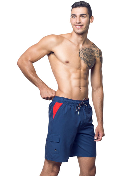 A man with short hair is standing against a white background, smiling. He is shirtless, displaying a tattoo on his left pectoral and shoulder, and is wearing BWET SWIMWEAR’s Neptune Beach Shorts in blue with red accents made from recycled material. One hand is on his waist, and the other rests by his side.