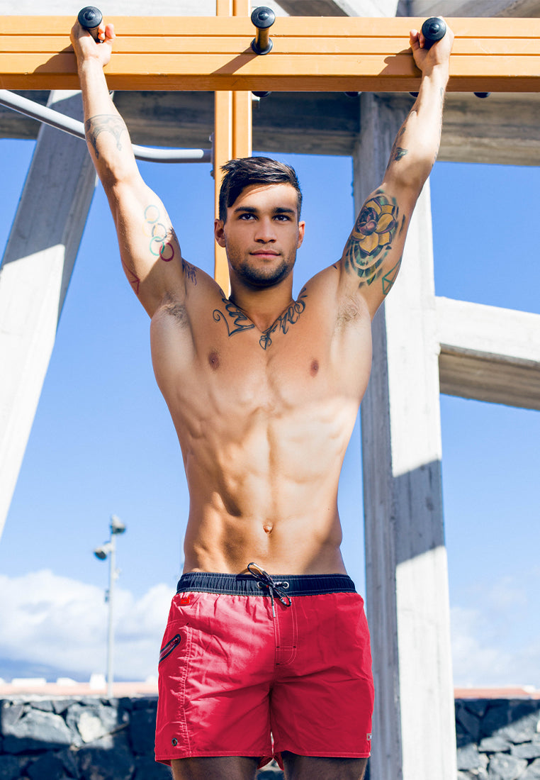 A shirtless man with tattoos on his arms and chest is hanging from an overhead exercise bar outdoors. He is wearing red BWET SWIMWEAR's Ozone Beach Shorts, made from recycled material, and looking straight ahead. The background includes a concrete structure and a clear blue sky.