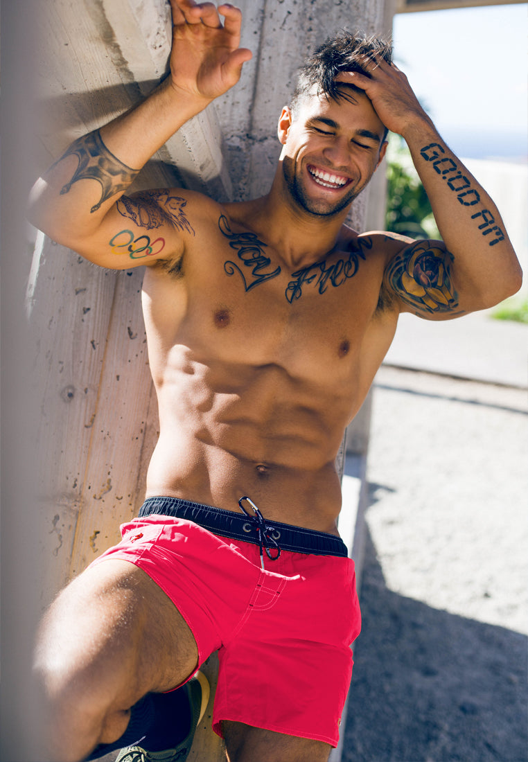 A man with dark hair and several tattoos on his arms and chest is smiling while leaning against a concrete structure. He is shirtless and wearing bright red BWET SWIMWEAR's Ozone Beach Shorts, known for their unbeatable style and functionality. The sun is shining, casting a warm glow on the scene.