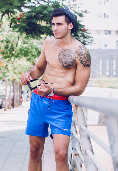 A man with a tattoo on his shoulder is leaning against a railing outdoors. He is shirtless, wearing BWET SWIMWEAR's Unbeatable Style and Functionality: Experience the Best with BWET Swimwear's Ozone Beach Shorts, a black hat, and holding sunglasses. Trees and buildings are visible in the background.