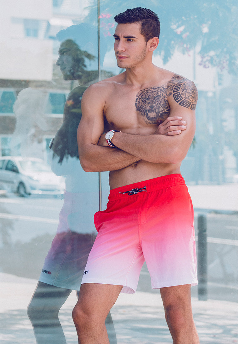 A shirtless man stands confidently by a reflective glass wall, donning BWET Swimwear's eco-friendly Sunrise Beach Shorts, distinguished by their striking red gradient quick-dry fabric. With his arms crossed and a tattoo on his left shoulder, he gazes into the distance. The backdrop features urban elements including cars, buildings, and trees, perfectly encapsulating contemporary beach fashion.