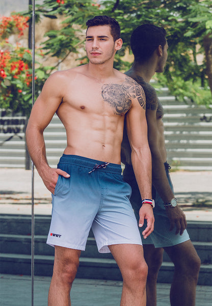 A man with a muscular physique stands shirtless outdoors, showcasing beach fashion. He wears BWET Swimwear's eco-friendly "Sunrise Beach Shorts" in a blue and white gradient, featuring the "wet" logo. Complementing his look is a red watch and a large tattoo on his left shoulder and chest. Steps and greenery are behind him, with his reflection visible.