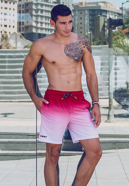 A muscular, young man with short dark hair is standing outdoors on a sunny day, leaning against a glass surface. He is shirtless, showing a large tattoo on his left shoulder and pectoral area, and wearing BWET Swimwear's eco-friendly Sunrise Beach Shorts from their Get Ready to Sizzle This Summer collection—a vibrant red-to-white gradient quick-dry design featuring a small logo.