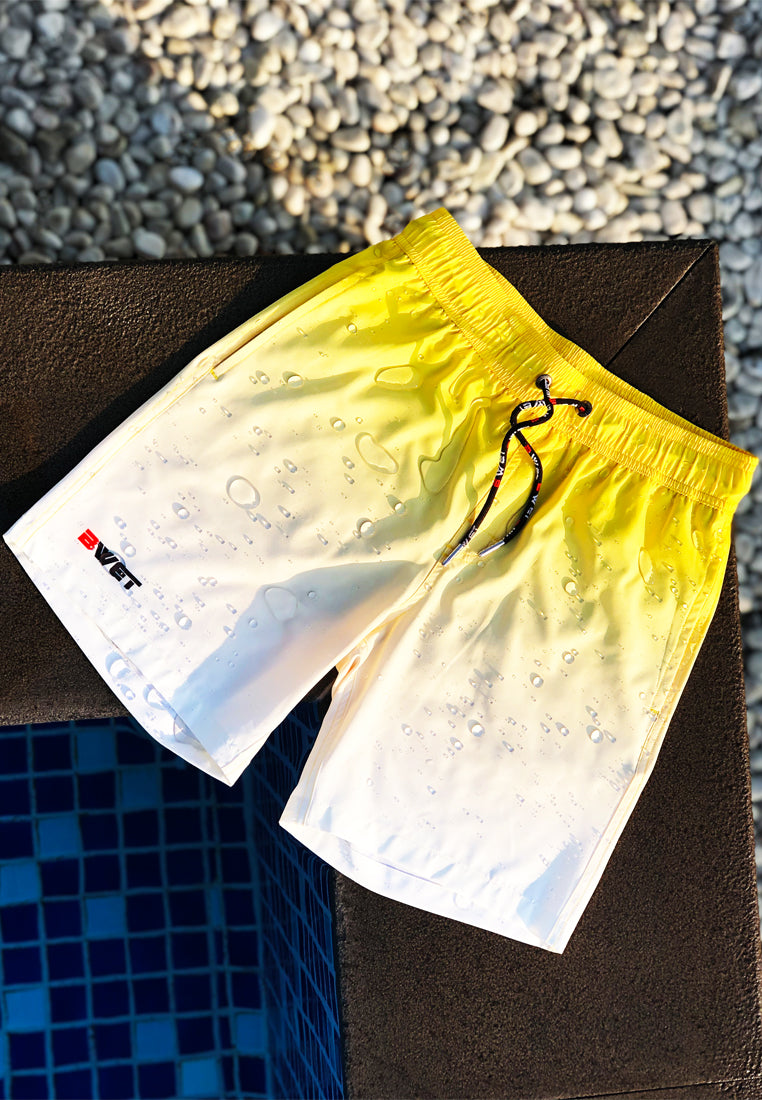 BWET Swimwear's Eco-Friendly Sunrise Beach Shorts, featuring a stylish yellow and white ombre design with a black drawstring, are placed on a brown border next to a swimming pool. The fabric, beautifully embellished with water droplets, complements the blue tiles of the pool and the surrounding white pebbles. This artistic setup highlights BWET Swimwear's Sunrise Beach Shorts as perfect beach essentials.