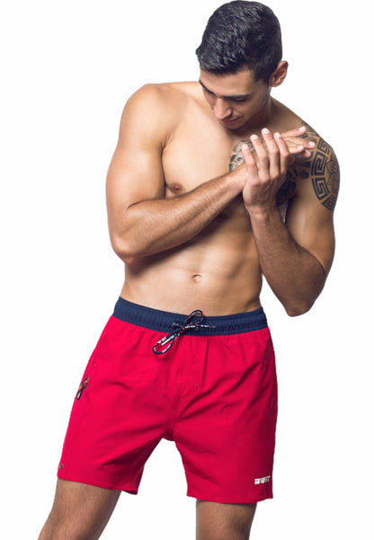 A shirtless young man with short hair looks down while holding his hands together. He has a tattoo on his upper right arm and is sporting BWET SWIMWEAR's Unbeatable Style and Functionality: Experience the Best with BWET Swimwear's Ozone Beach Shorts in bright red with a black waistband, crafted from recycled materials. The background is plain white.