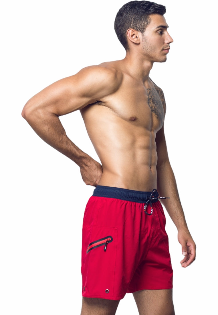 A shirtless man stands in profile, wearing bright red Ozone Beach Shorts from BWET SWIMWEAR that feature a black waistband and drawstrings. His left hand rests on his left hip, showcasing a tattoo on his chest and shoulder against the plain white background. Made from recycled material, these shorts are part of BWET Swimwear's "Unbeatable Style and Functionality" collection.