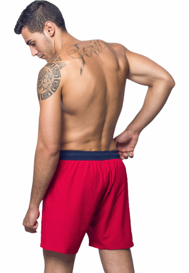 A man stands turned slightly away, showcasing his back and arm tattoos. He is shirtless and wears BWET SWIMWEAR's Ozone Beach Shorts in red, featuring a black waistband and crafted from recycled material. His right hand rests behind his back as he looks down to his left against a white background. Experience unbeatable style and functionality with BWET Swimwear's Ozone Beach Shorts.