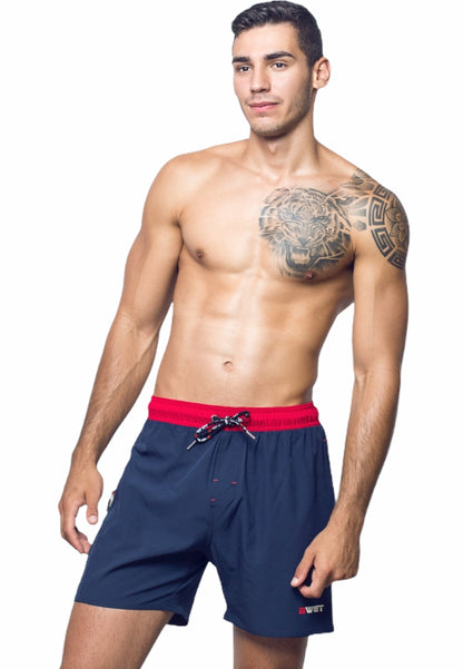 A man stands posing against a white background. He is shirtless, revealing a tattoo of a tiger on his upper left chest and shoulder. He is wearing the Unbeatable Style and Functionality: Experience the Best with BWET Swimwear's Ozone Beach Shorts from BWET SWIMWEAR in navy blue with a red waistband and a small white and red logo near the right hem.