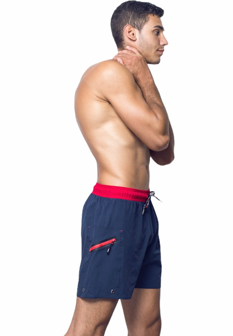 A man with short dark hair stands in profile against a white background. He is shirtless and wearing BWET SWIMWEAR's Ozone Beach Shorts, which feature a navy blue color with a red waistband and a red zipper pocket made from recycled material. His left hand is touching his neck while his right arm hangs by his side.