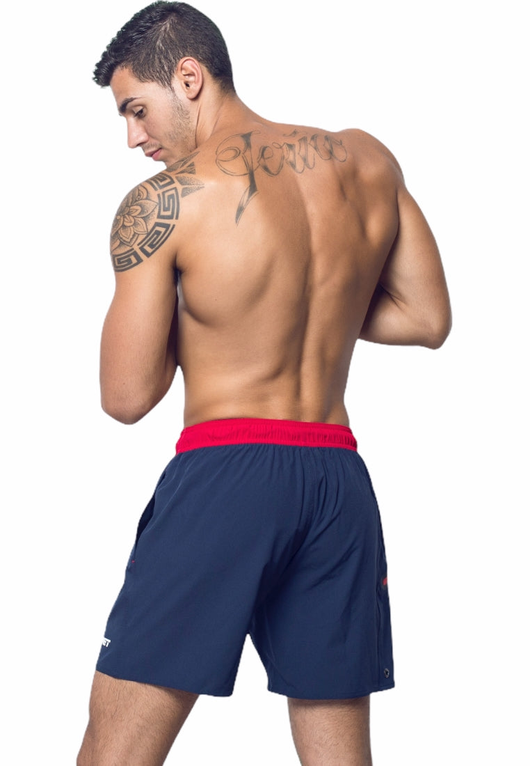 A muscular man with a tattoo on his upper back and left shoulder stands shirtless, showcasing BWET SWIMWEAR's Ozone Beach Shorts in navy blue with a red waistband. His body is turned slightly, revealing his back and part of his left arm.