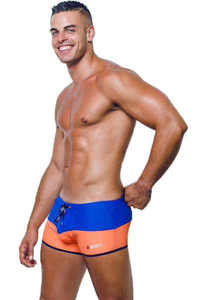 A smiling, muscular man with short hair is modeling BWET Swimwear's high-quality and stylish Sentosa Beach Trunks in blue and orange. The swimwear features a drawstring, a small logo on the left leg, and offers UV protection. He is posing confidently against a plain white background.