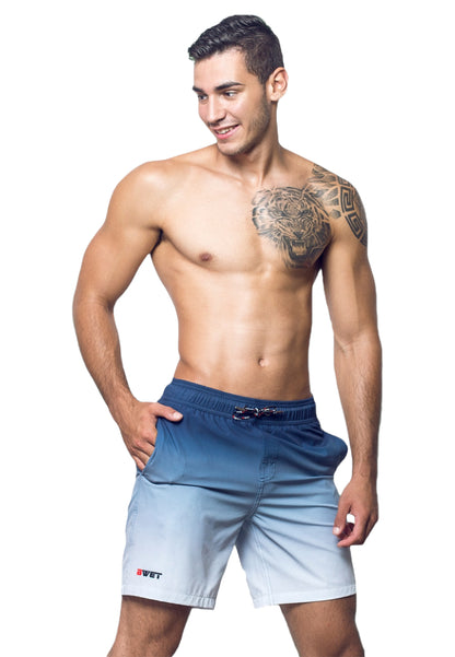 A young man stands smiling, wearing BWET Swimwear's Eco-Friendly Sunrise Beach Shorts in a light blue and navy gradient with his left hand in the pocket. His right arm rests by his side, revealing a detailed bear tattoo on his left shoulder and chest. He is shirtless and appears to be in a relaxed pose, epitomizing beach fashion as he gets ready to sizzle this summer.