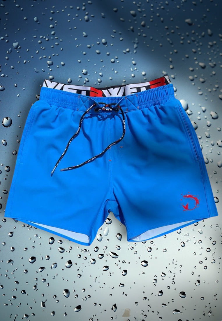 A pair of bright blue "Be Extraordinary at the Beach with Infinity Beach Shorts" by BWET Swimwear, featuring a black adjustable drawstring, displayed on a surface with water droplets. Made from eco-friendly materials, the waistband boasts a distinct black and red design, highlighted by a small red fish logo on the front left leg.