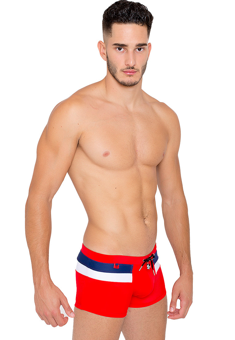 A man with short, dark hair is donning the Discover "Rooftop" by BWET Swimwear: Comfortable and Sporty Beach Trunks. The red swim trunks feature a stylish white and navy blue horizontal stripe. Standing sideways with a serious expression directed at the camera, his left arm rests by his side, showcasing his fit, muscular physique. Thanks to the quick-dry material of BWET SWIMWEAR's trunks, he remains comfortable all day long.