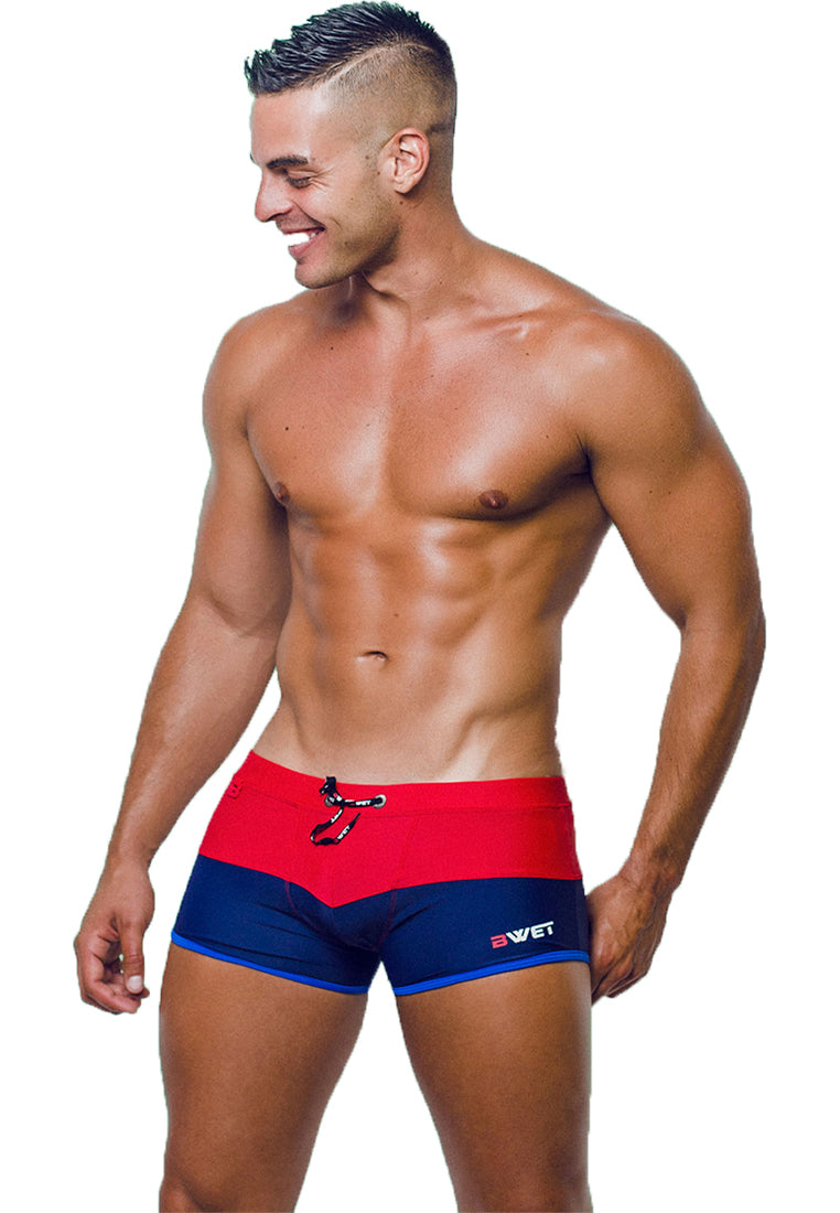 A muscular man with a short haircut is standing and smiling, looking towards his left. He is wearing a pair of Sentosa Beach Trunks from BWET Swimwear, predominantly red with blue accents and a drawstring at the waist. These trunks offer ultra-chlorine resistance and UV protection, with the brand logo "BWET" prominently displayed on the left side.