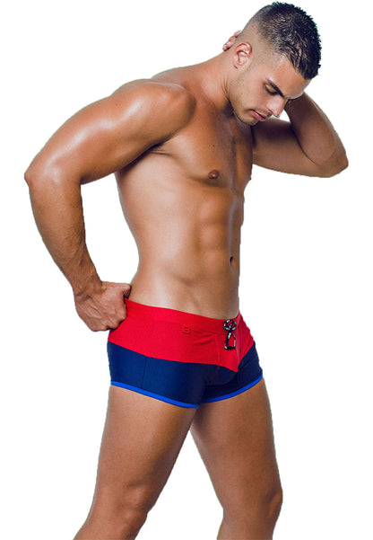 A muscular man with short hair stands against a white background, looking downward. He is wearing form-fitting Sentosa Beach Trunks from BWET Swimwear. These high-quality and stylish swimwear trunks are red at the waist and dark blue at the legs, featuring a decorative buckle at the front. They offer UV protection and ultra-chlorine resistance.