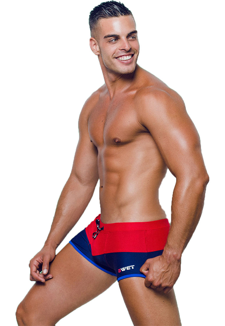 A muscular man with short hair smiles and glances to the side while posing in red and blue Sentosa Beach Trunks by BWET Swimwear. The plain white background accentuates his athletic physique and the high-quality, stylish swimwear, which offers UV protection and ultra-chlorine resistance for lasting durability.