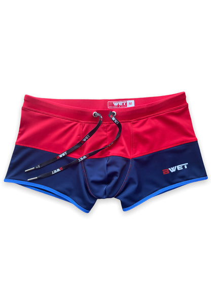 The red and navy blue Sentosa Beach Trunks by BWET Swimwear feature a drawstring waistband and a white "BWET" logo on the lower right side. These high-quality and stylish swim trunks boast a contrasting color block design, close-fitting style, UV protection, and ultra-chlorine resistance for ultimate durability.